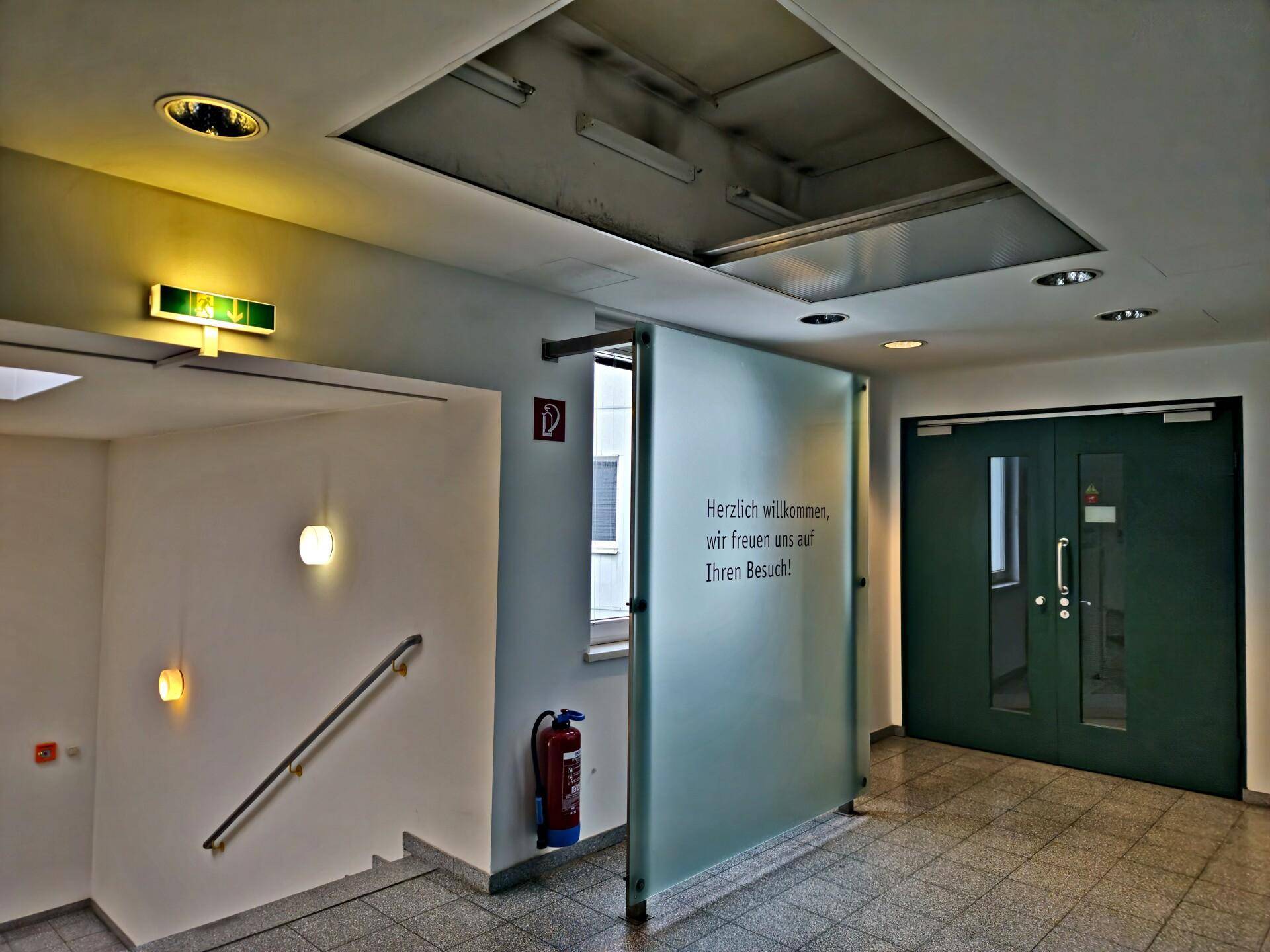 Eingangsbereich mit Treppe, matter Glastür und Sicherheitshinweisen an den Wänden.