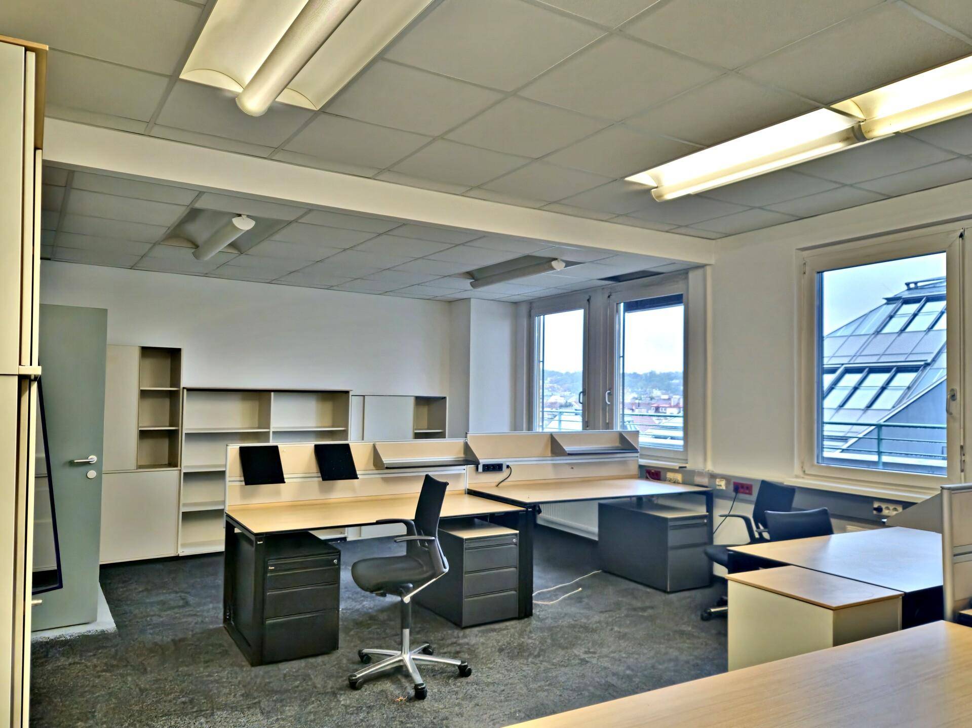 Geräumiges Büro mit mehreren Arbeitsplätzen, Regalen und Blick auf die Stadt durch große Fenster.