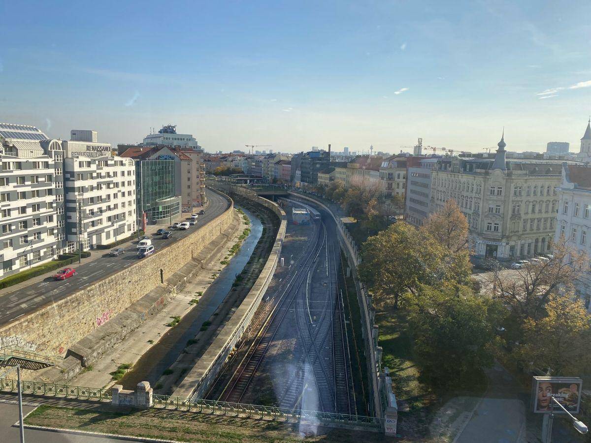 Weitläufiger Blick auf eine Stadtlandschaft mit Fluss, Bahngleisen und Gebäuden unter blauem Himmel.
