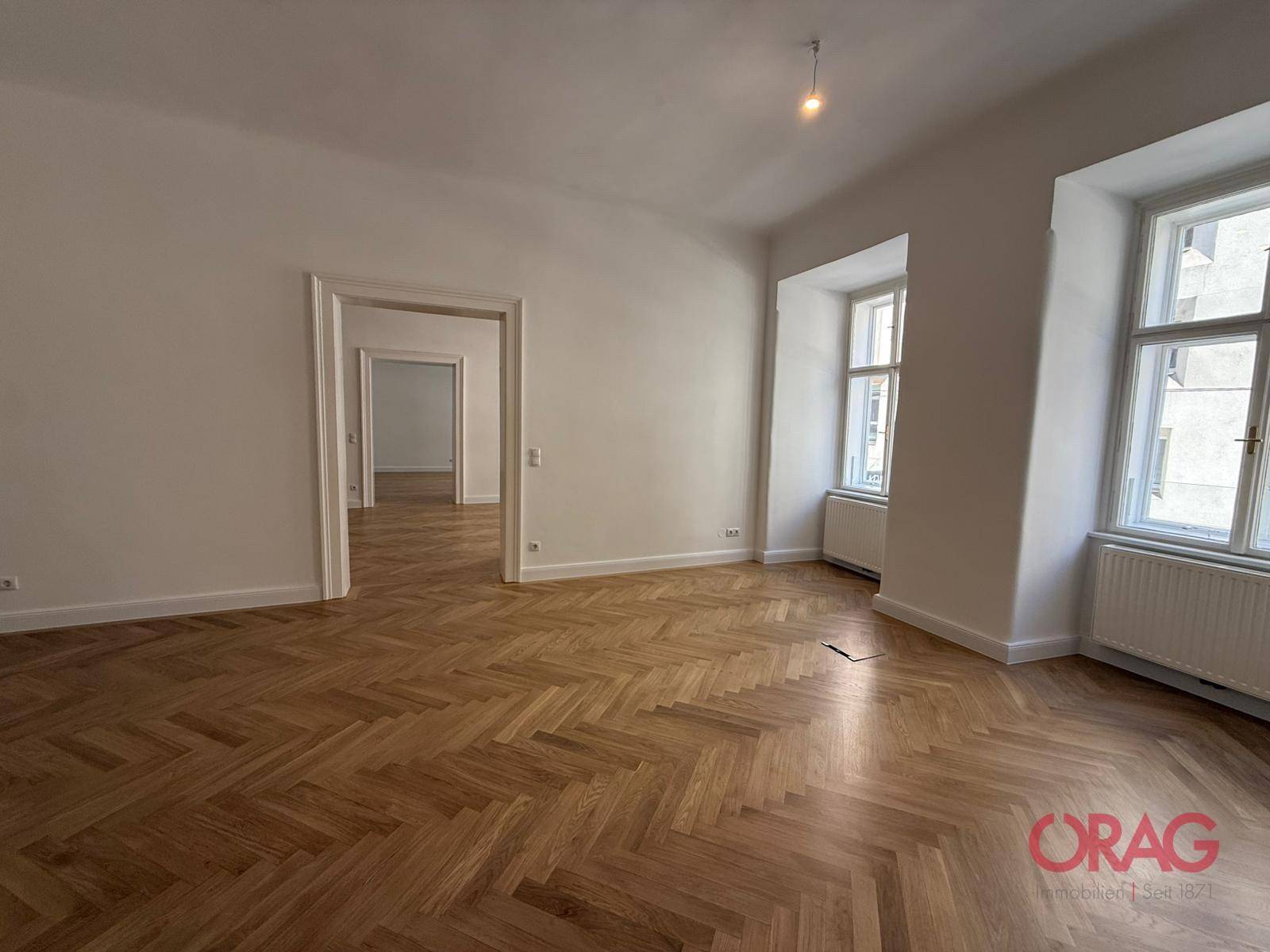 Geräumiges Zimmer mit klassischem Fischgrätparkett und zwei großen Fenstern, die viel Tageslicht hereinlassen.