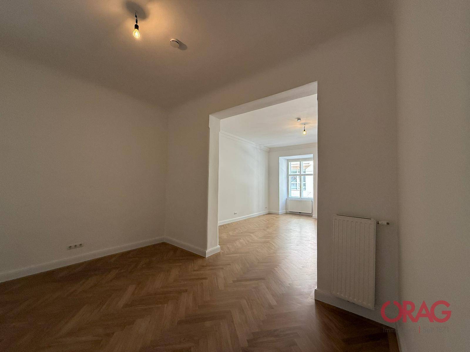 Heller Raum mit elegantem Fischgrätparkett und offenem Durchgang zu einem weiteren Zimmer, ideal für flexible Nutzung.