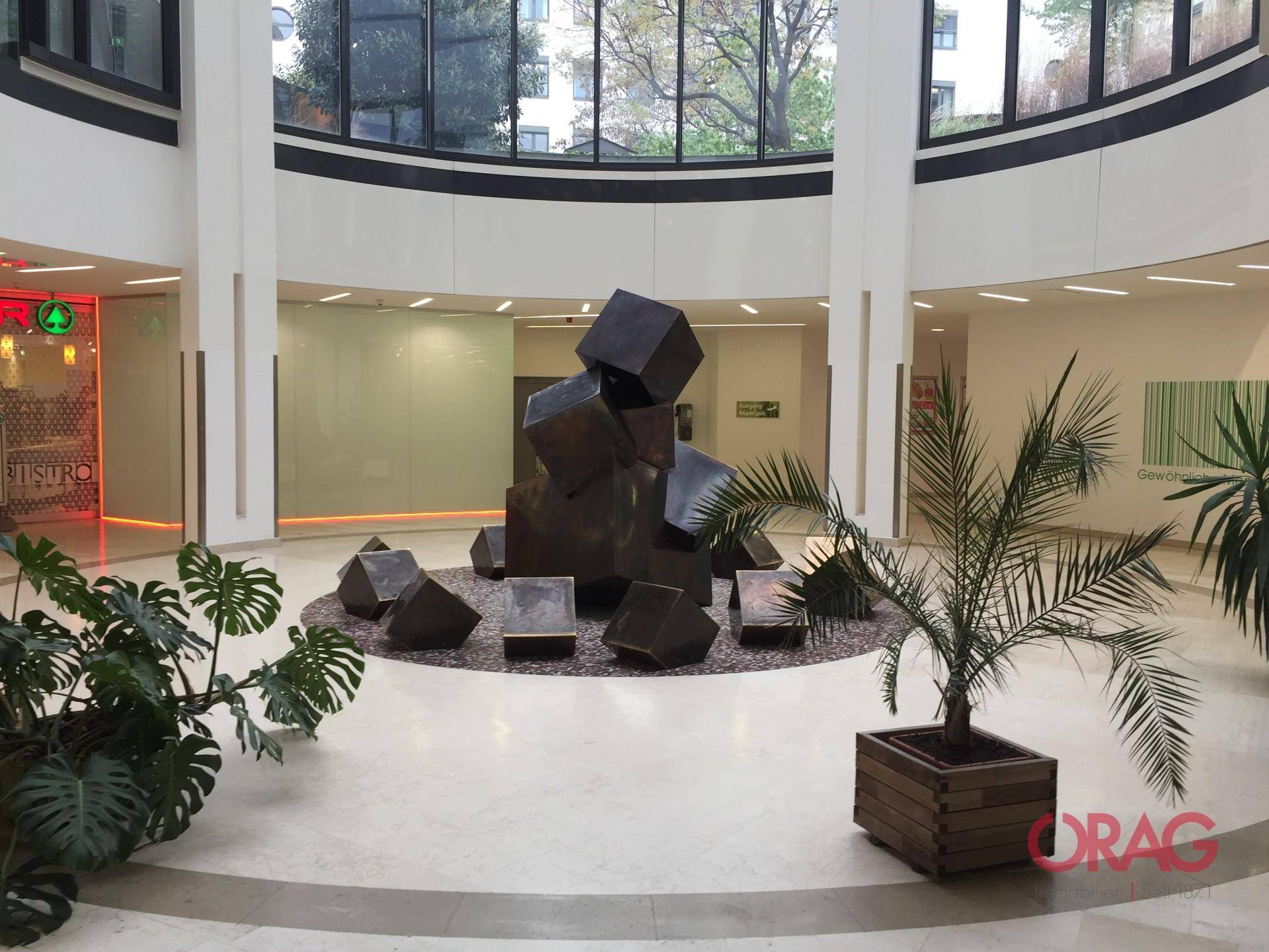 Großzügiges, helles Atrium mit moderner Skulptur, Pflanzen und glänzendem Fliesenboden.