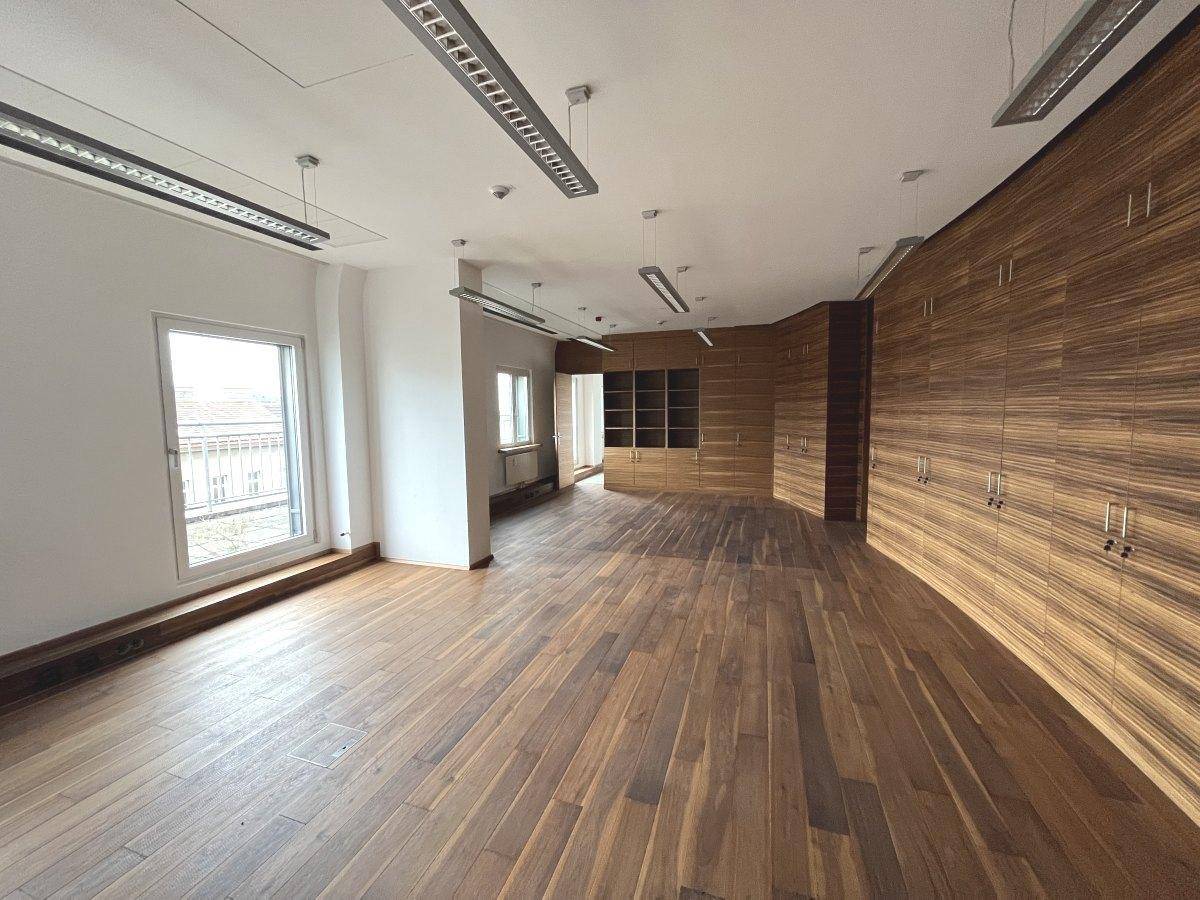 Geräumiges, leeres Büro mit Holzboden, großen Fenstern und einer Wandverkleidung aus dunklem Holz.