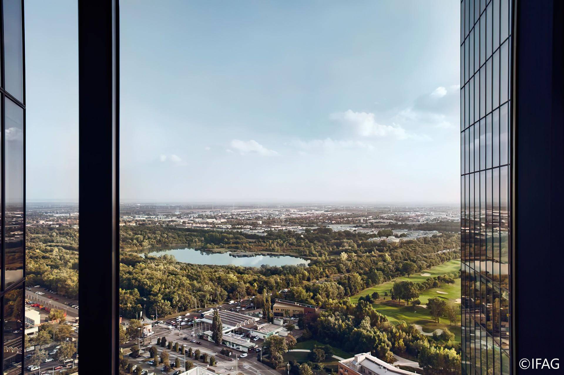 Weitläufiger Ausblick aus einem Bürofenster auf eine grüne Landschaft mit See und Stadt.