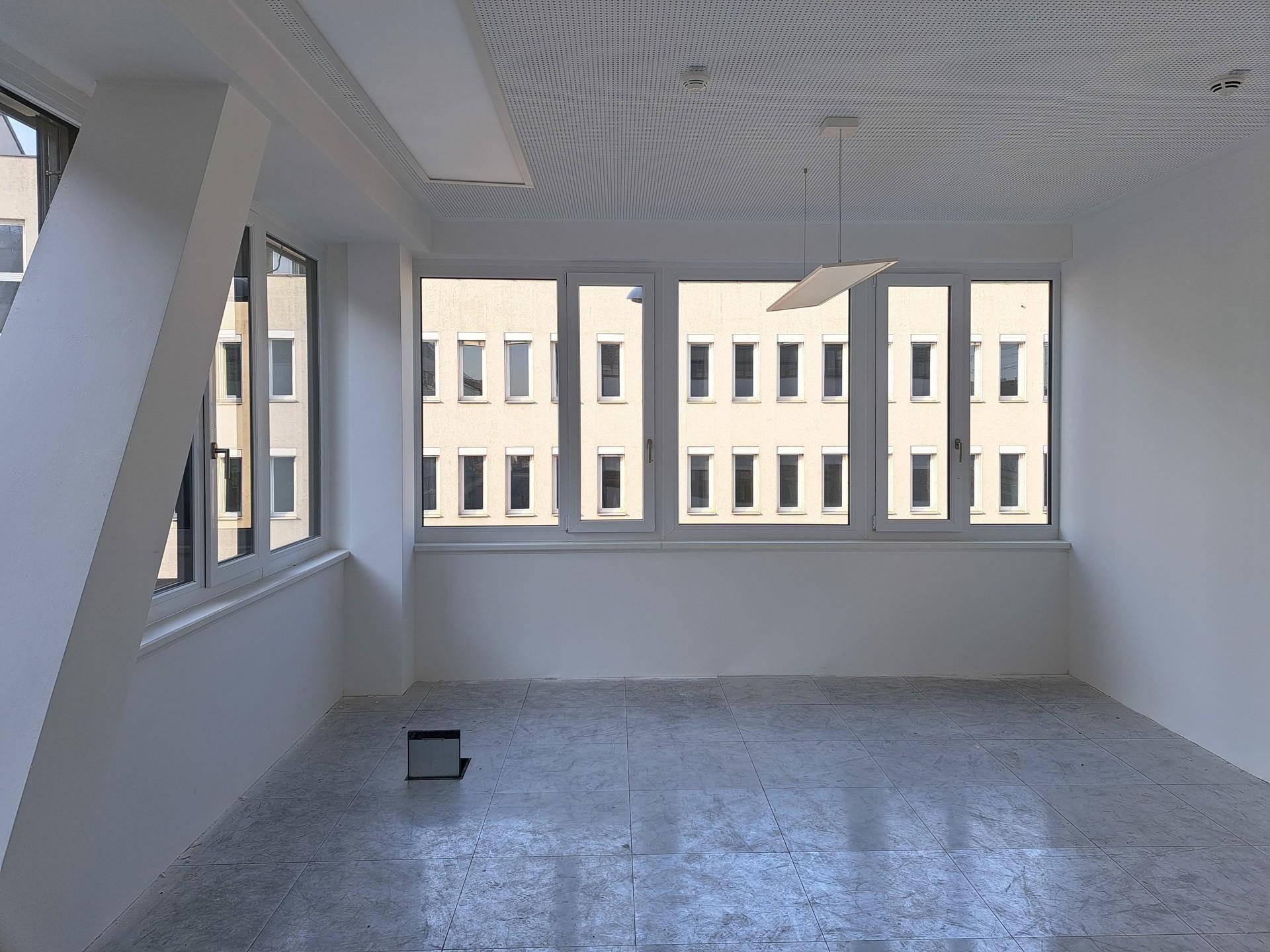 Leerer Büroraum mit großen Fenstern, die den Blick auf ein gegenüberliegendes Gebäude freigeben.
