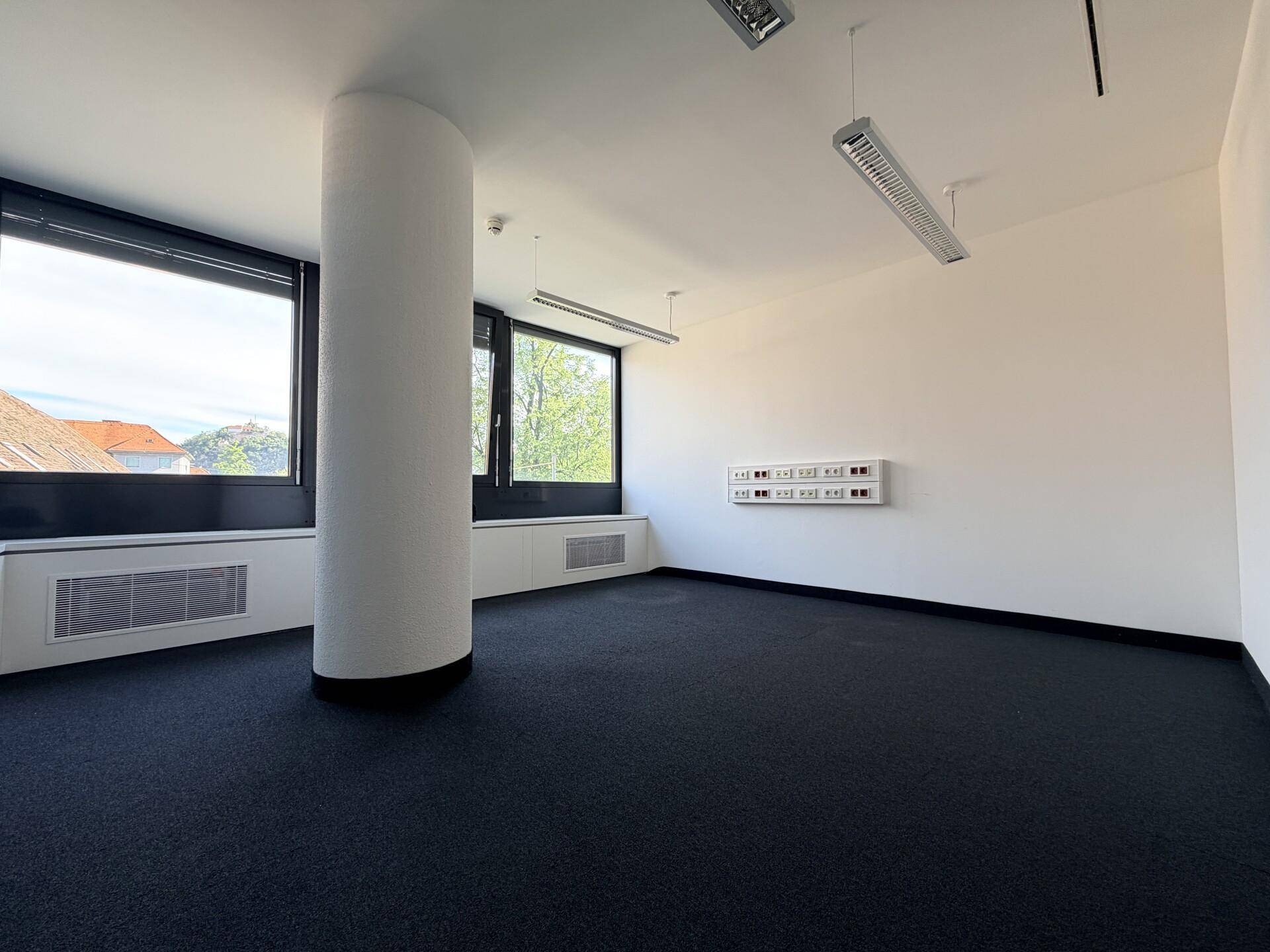 Geräumiges Büro mit dunklem Teppichboden, großen Fenstern und einer zentralen Säule.