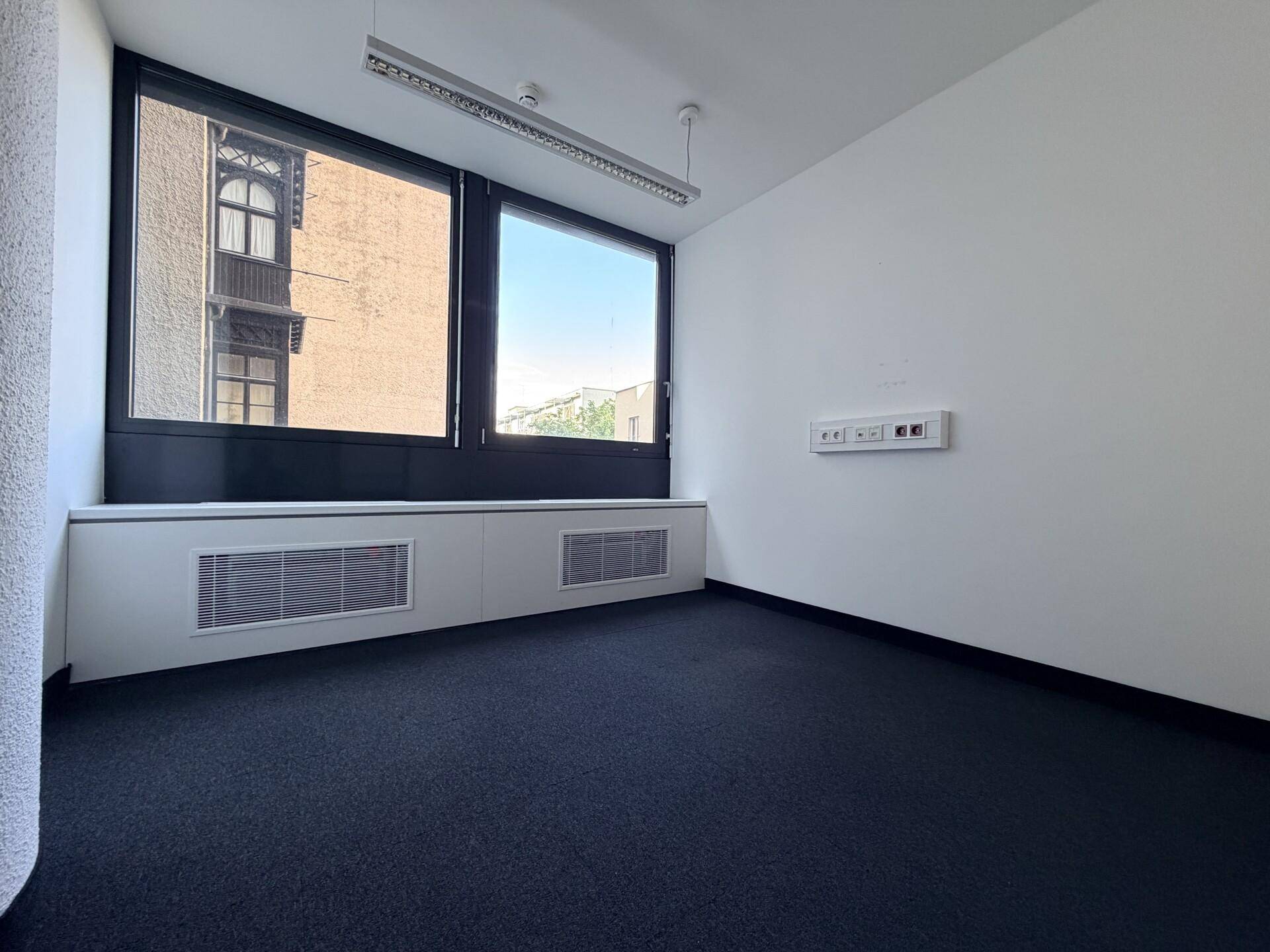 Helles Büro mit großen Fenstern, die einen Blick auf die Nachbargebäude ermöglichen, und dunklem Teppichboden.