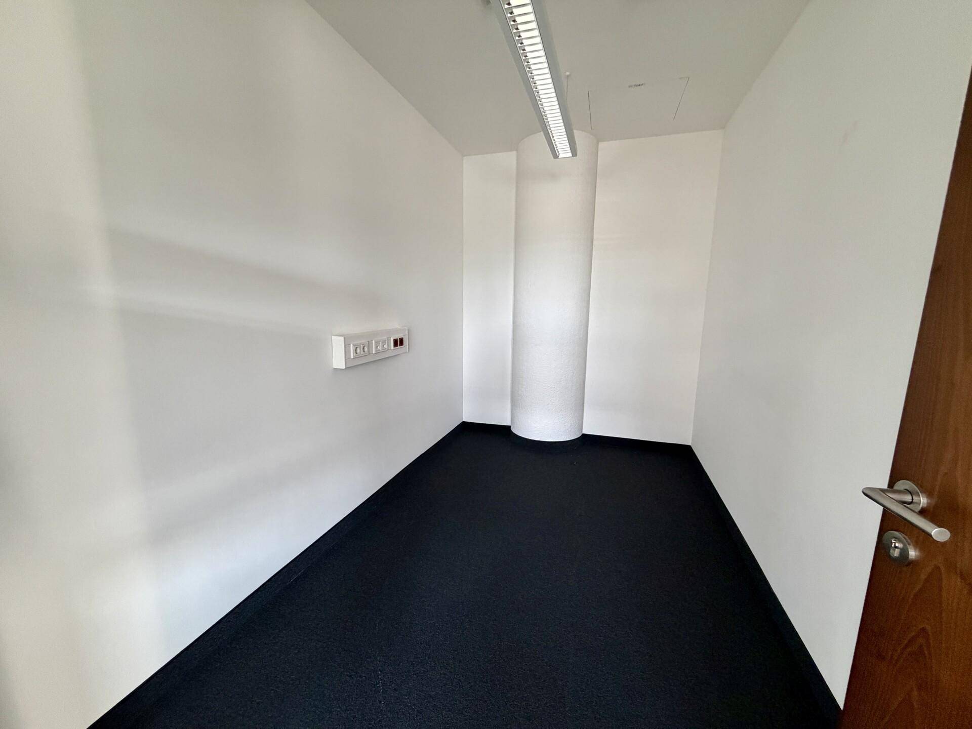 Kleines, leeres Büro mit dunklem Teppichboden, weißen Wänden und einer runden Säule im Eckbereich.