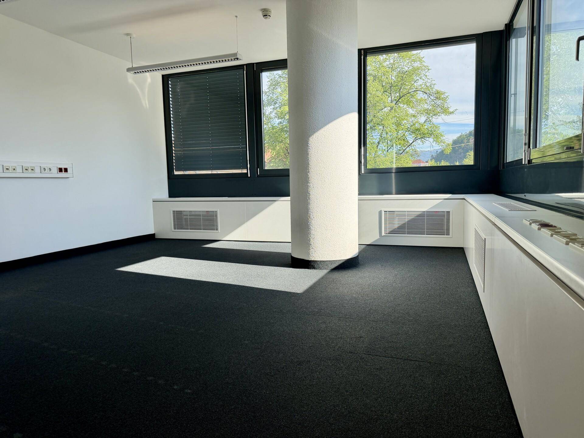 Helles, leeres Büro mit dunklem Teppichboden, großen Fenstern und einer runden Säule im Raum.