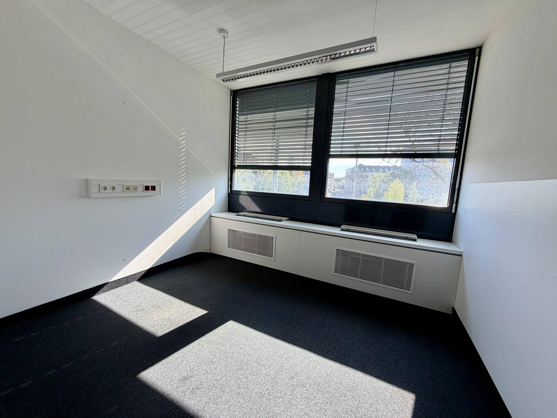 Leeres Büro mit Teppichboden, großen Fenstern mit Jalousien und Sonnenlicht, das Streifen auf den Boden wirft.