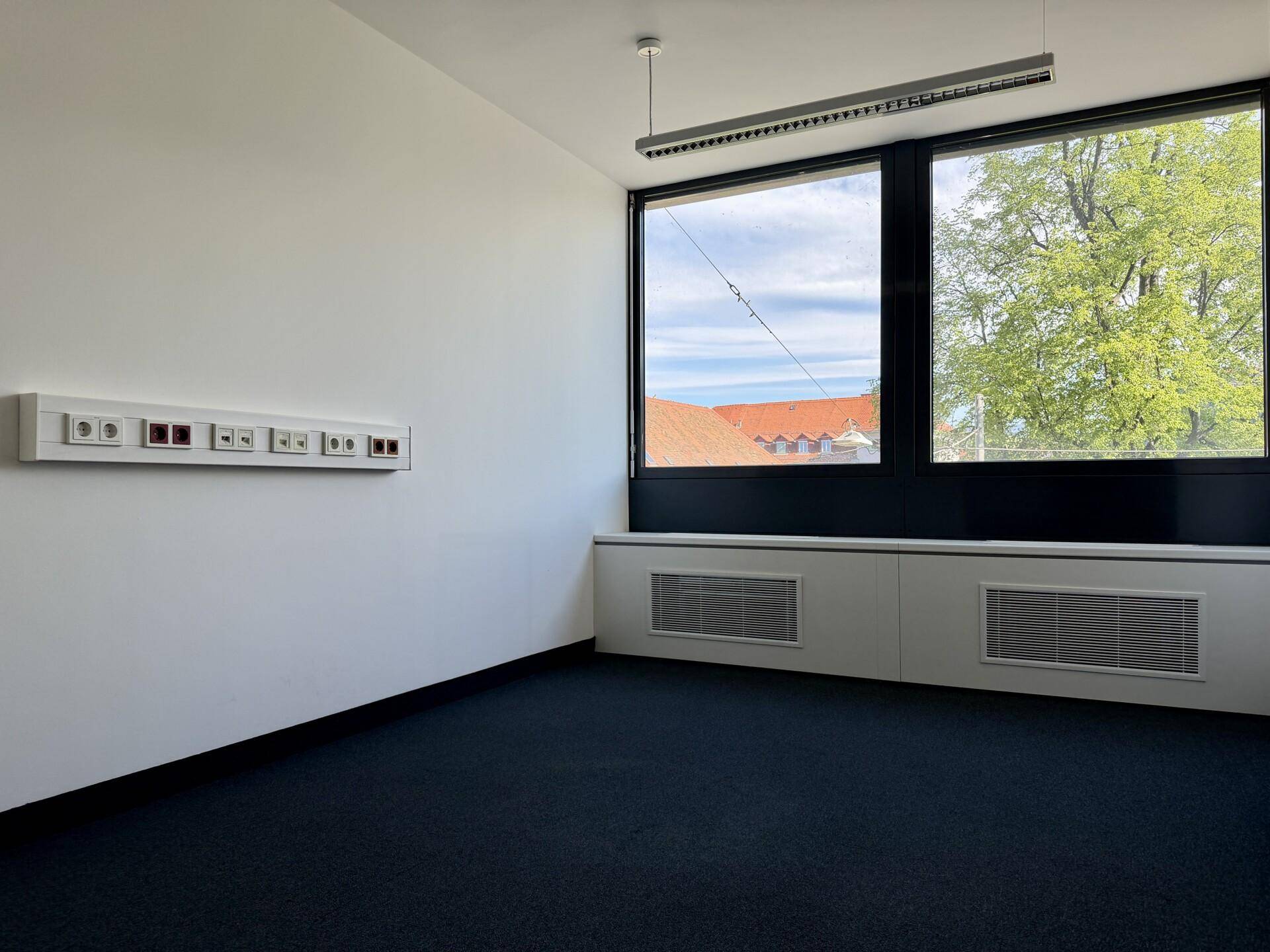 Leeres Büro mit Teppichboden, großen Fenstern und Blick auf Dächer und den Himmel, gut beleuchtet.