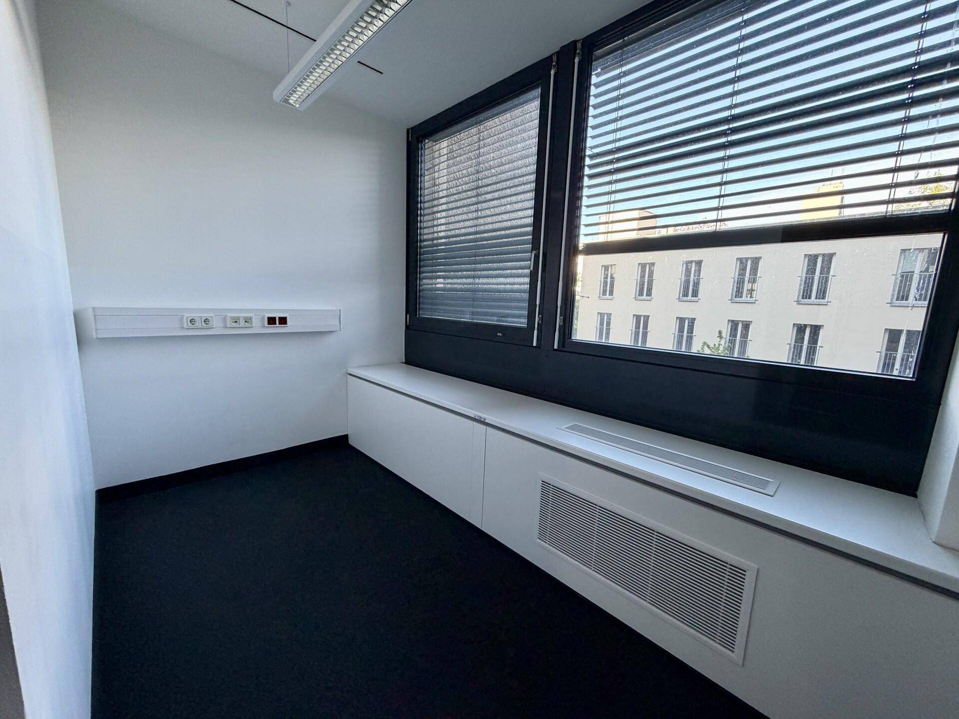 Modernes Büro mit Teppichboden, großen Fenstern mit Jalousien und einem weiten Blick auf die Stadt.