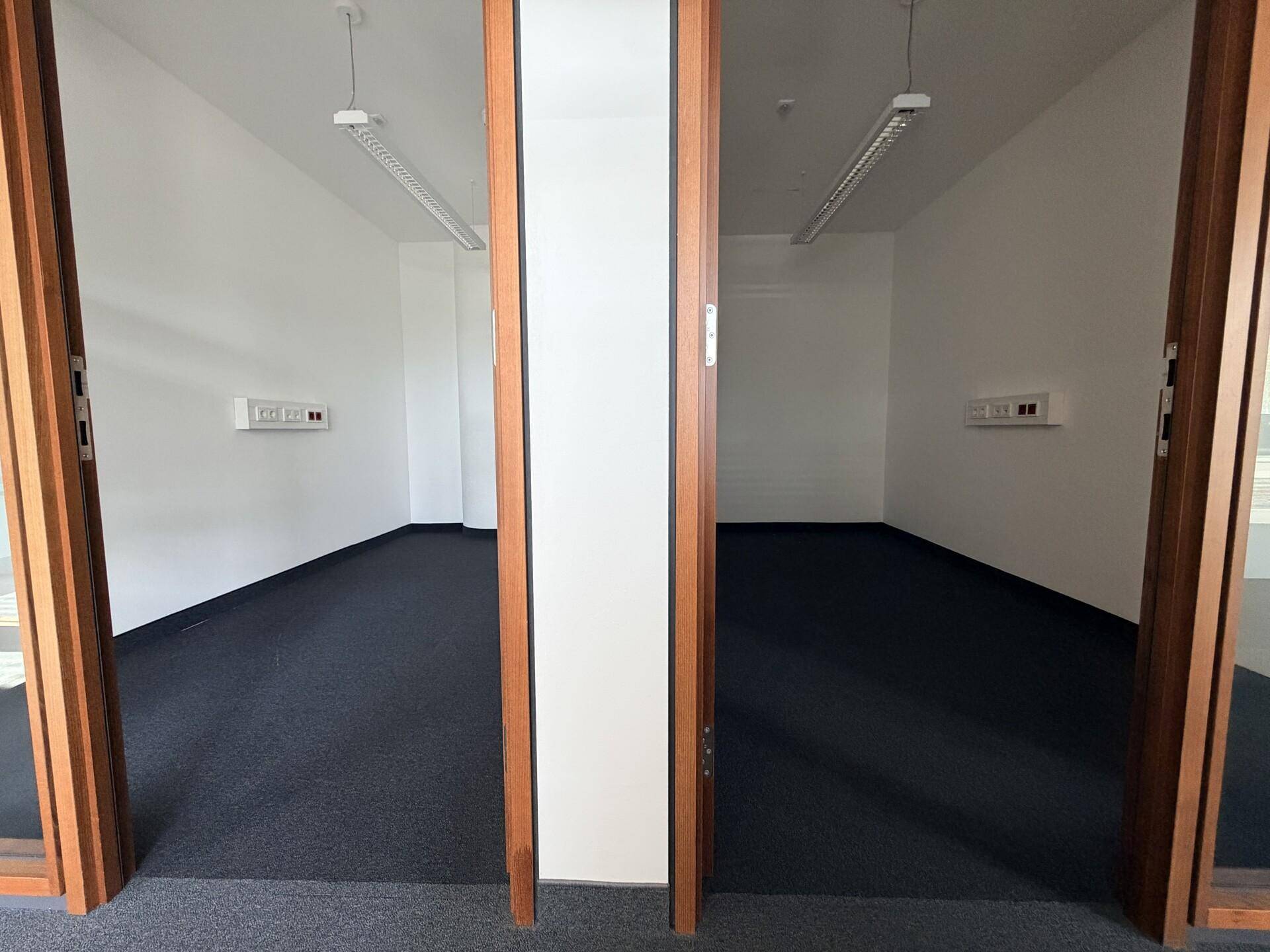 Zwei leere Büroräume mit Teppichboden und weißen Wänden, durch offene Holztüren sichtbar und gut beleuchtet.