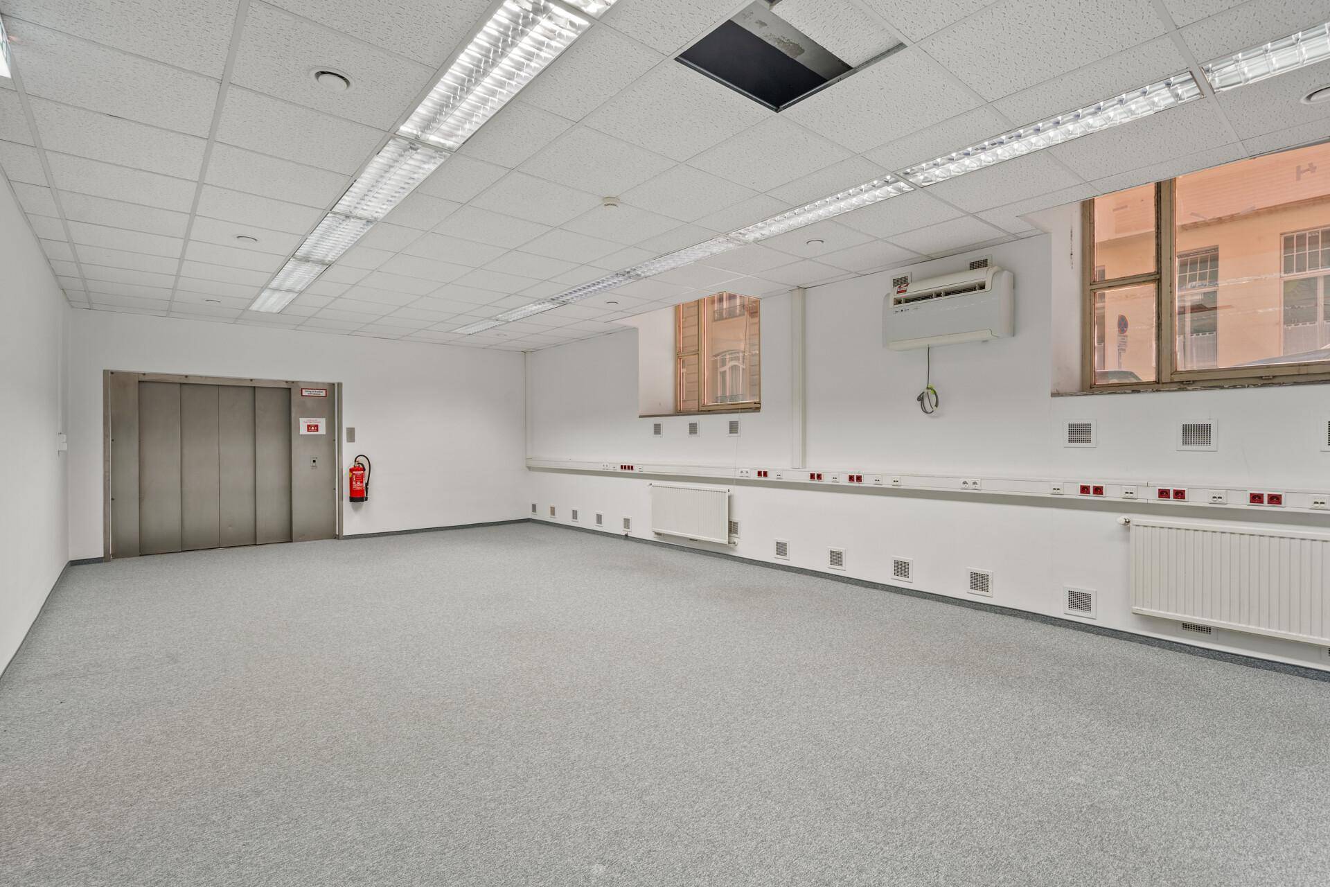 Helles Büro mit Teppichboden, Aufzugzugang und Heizkörpern unter den Fenstern, bereit zur Nutzung.