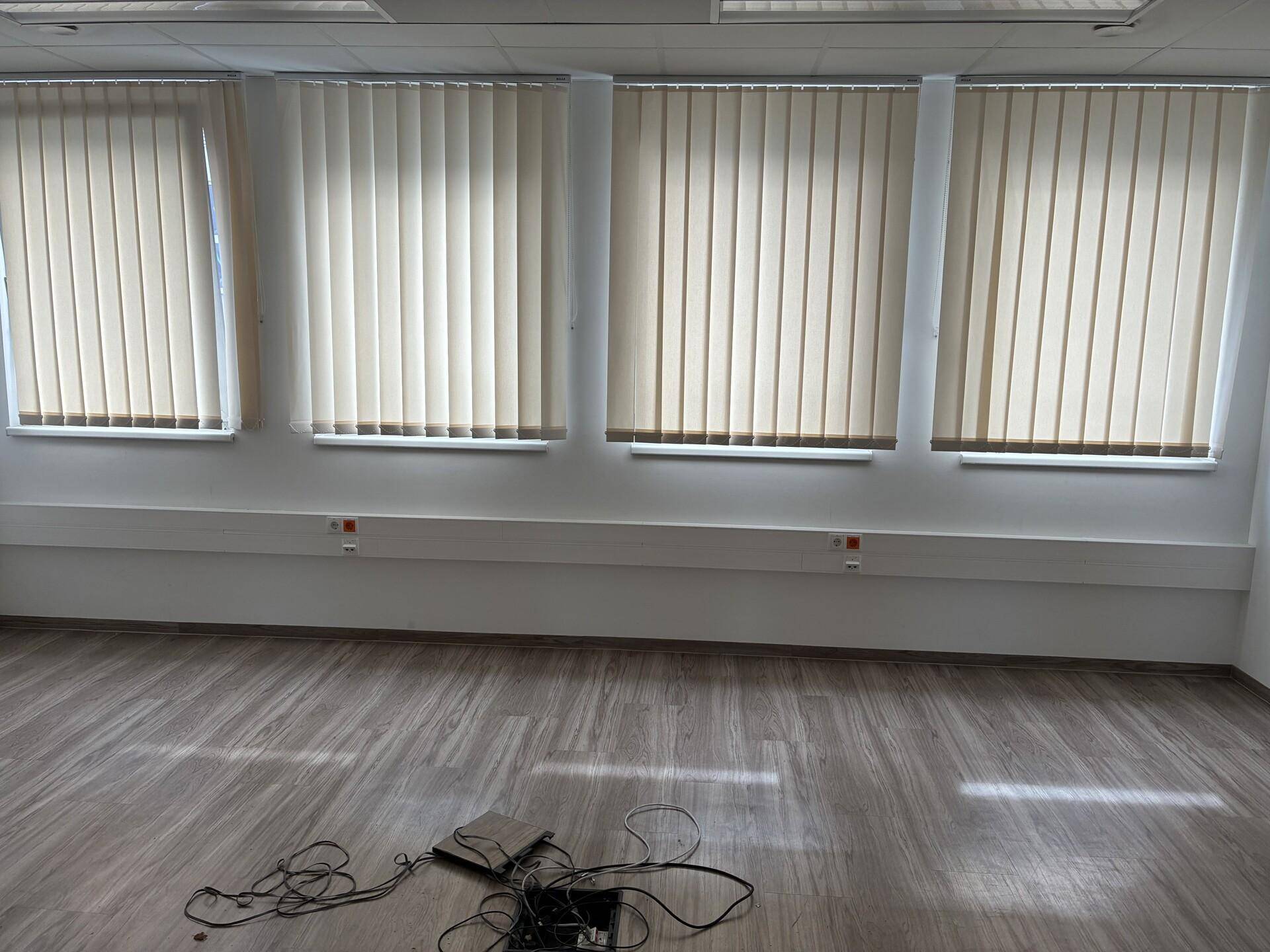 Leerer Büroraum mit hellem Laminatboden und vertikalen Jalousien an den Fenstern.