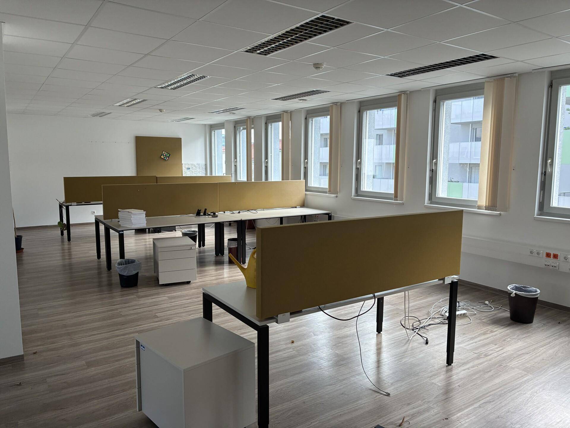 Großraumbüro mit mehreren Arbeitsplätzen, Trennwänden und großen Fenstern.