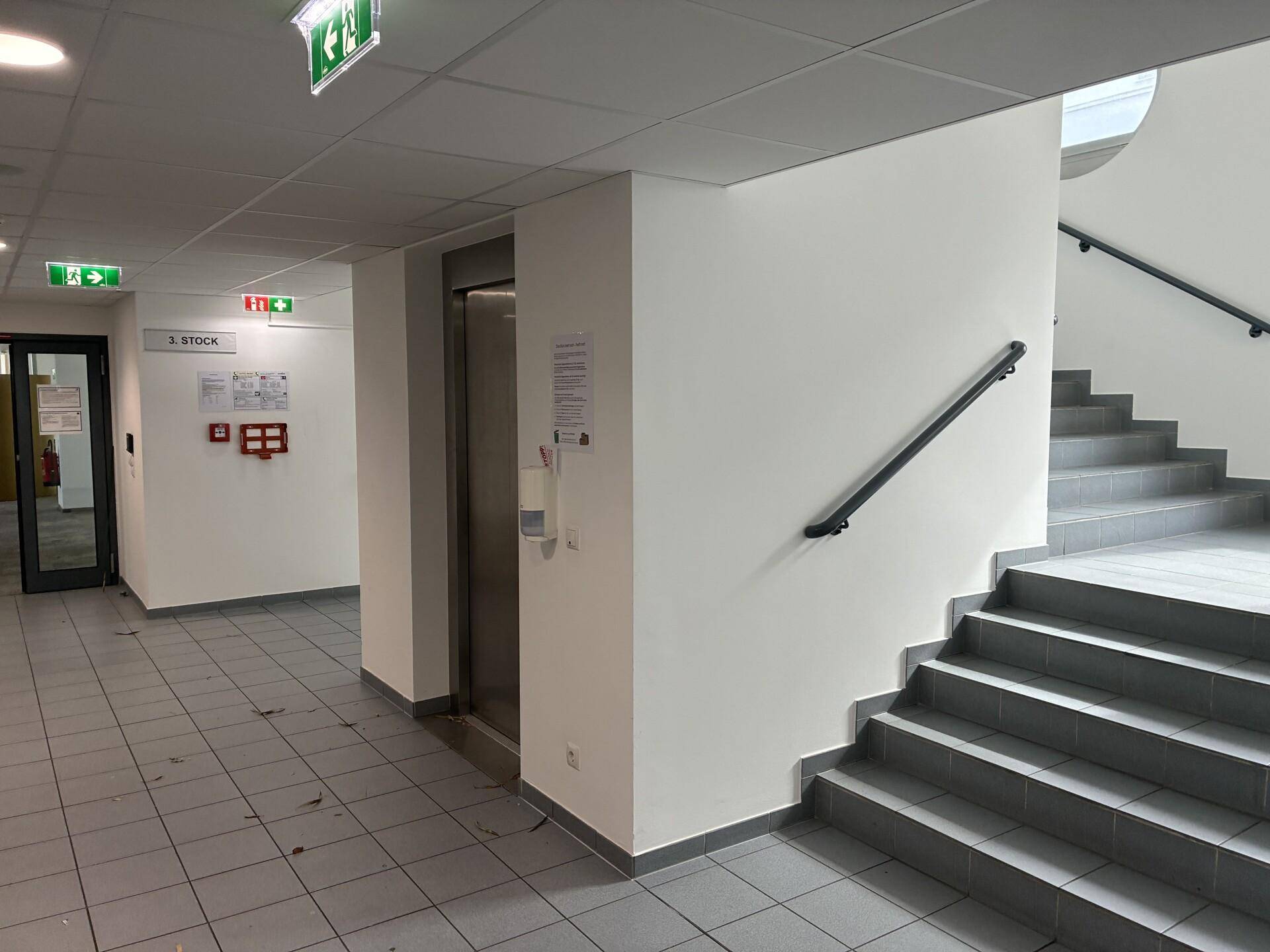 Heller Flur mit Treppenhaus, Aufzug und Hinweisschildern im Bürogebäude.