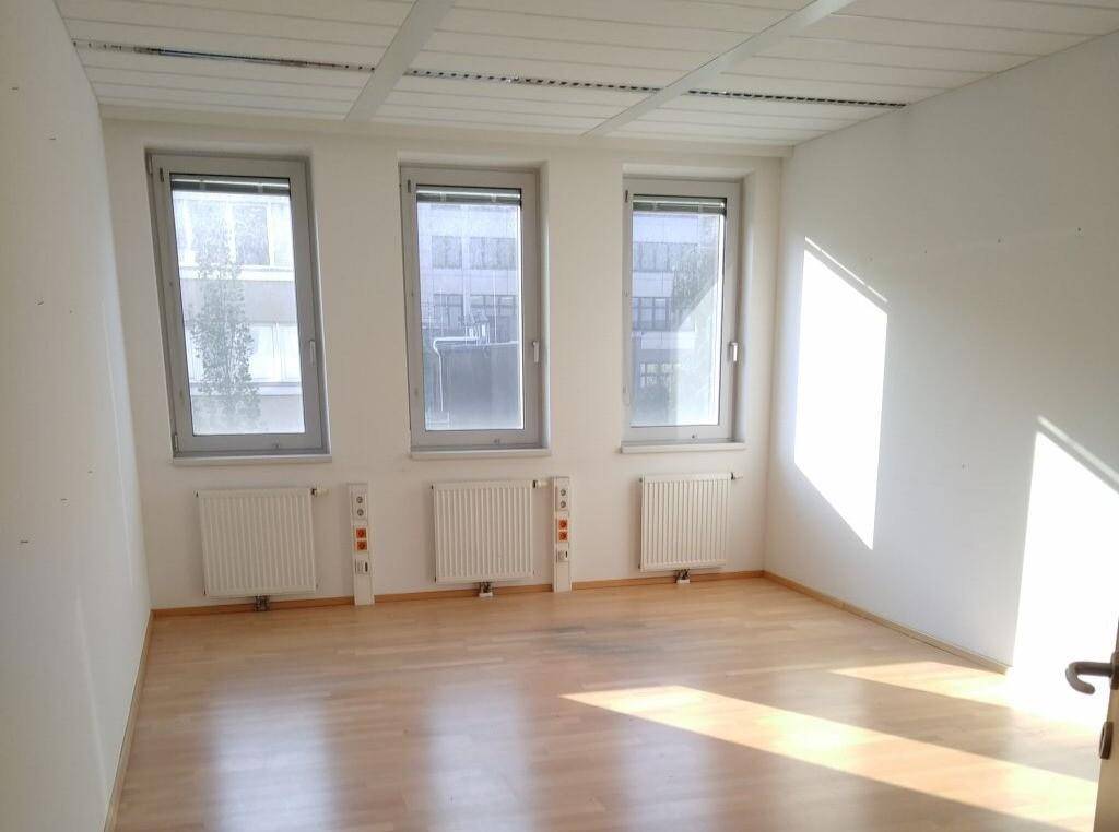 Heller Büroraum mit drei Fenstern, hellem Laminatboden und Heizkörpern unter einer Rasterdecke.
