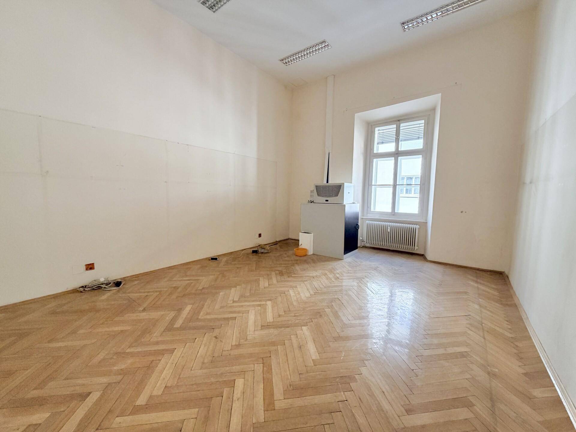 Geräumiges Büro mit Fischgrätparkett, hohen Decken und einem großen Fenster.