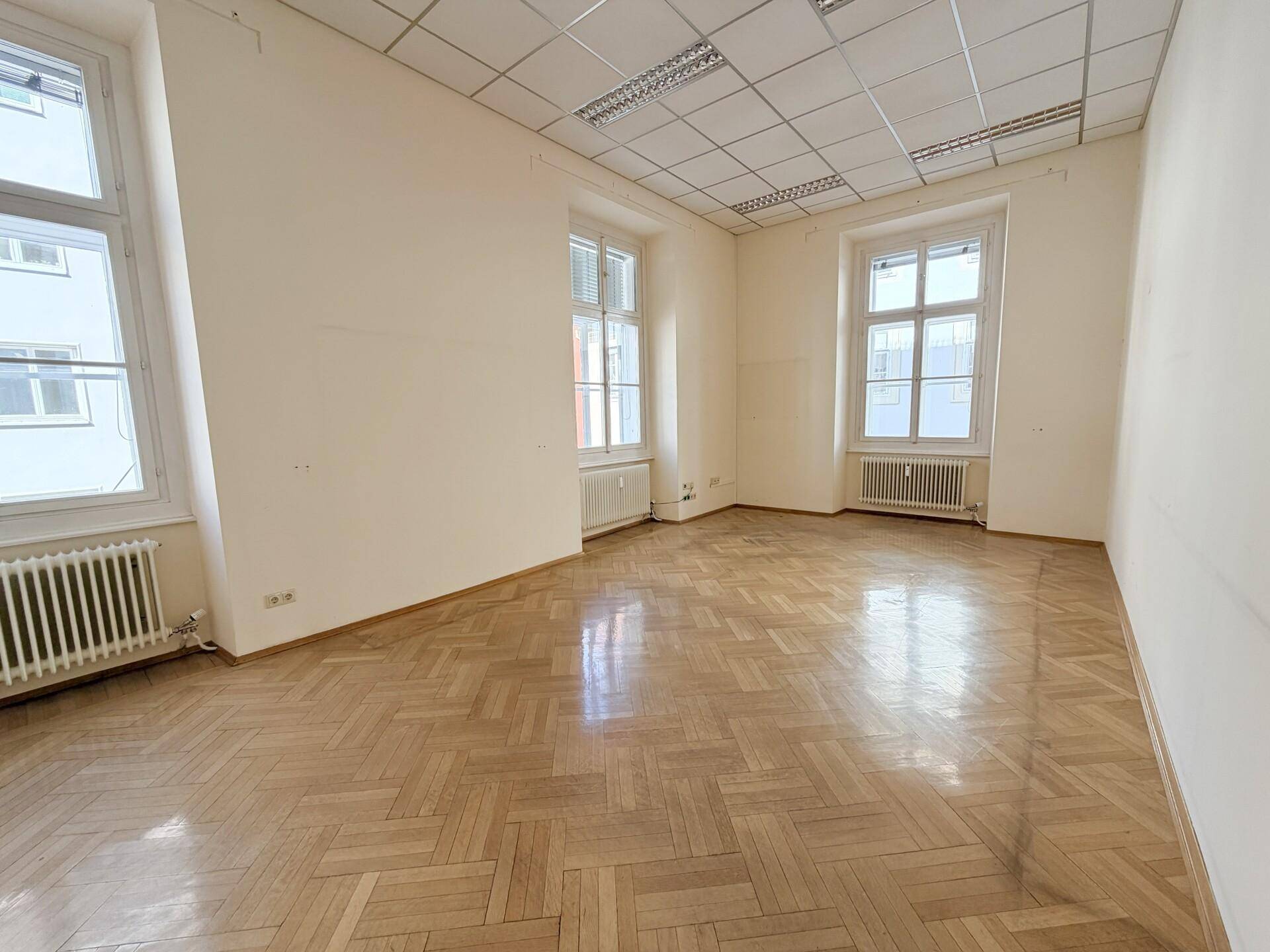 Großes Büro mit zwei Fenstern, Fischgrätparkett und abgehängter Decke.