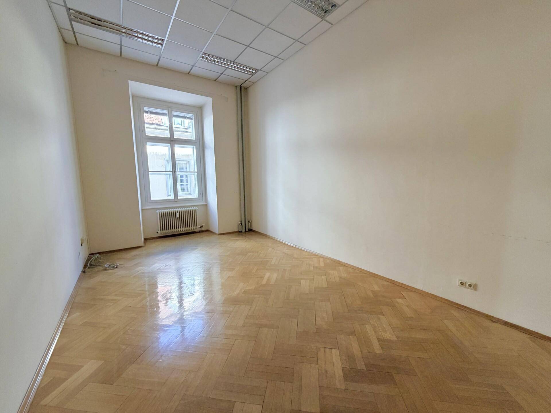 Helles Büro mit Fischgrätparkett, abgehängter Decke und einem Fenster mit Heizkörper.