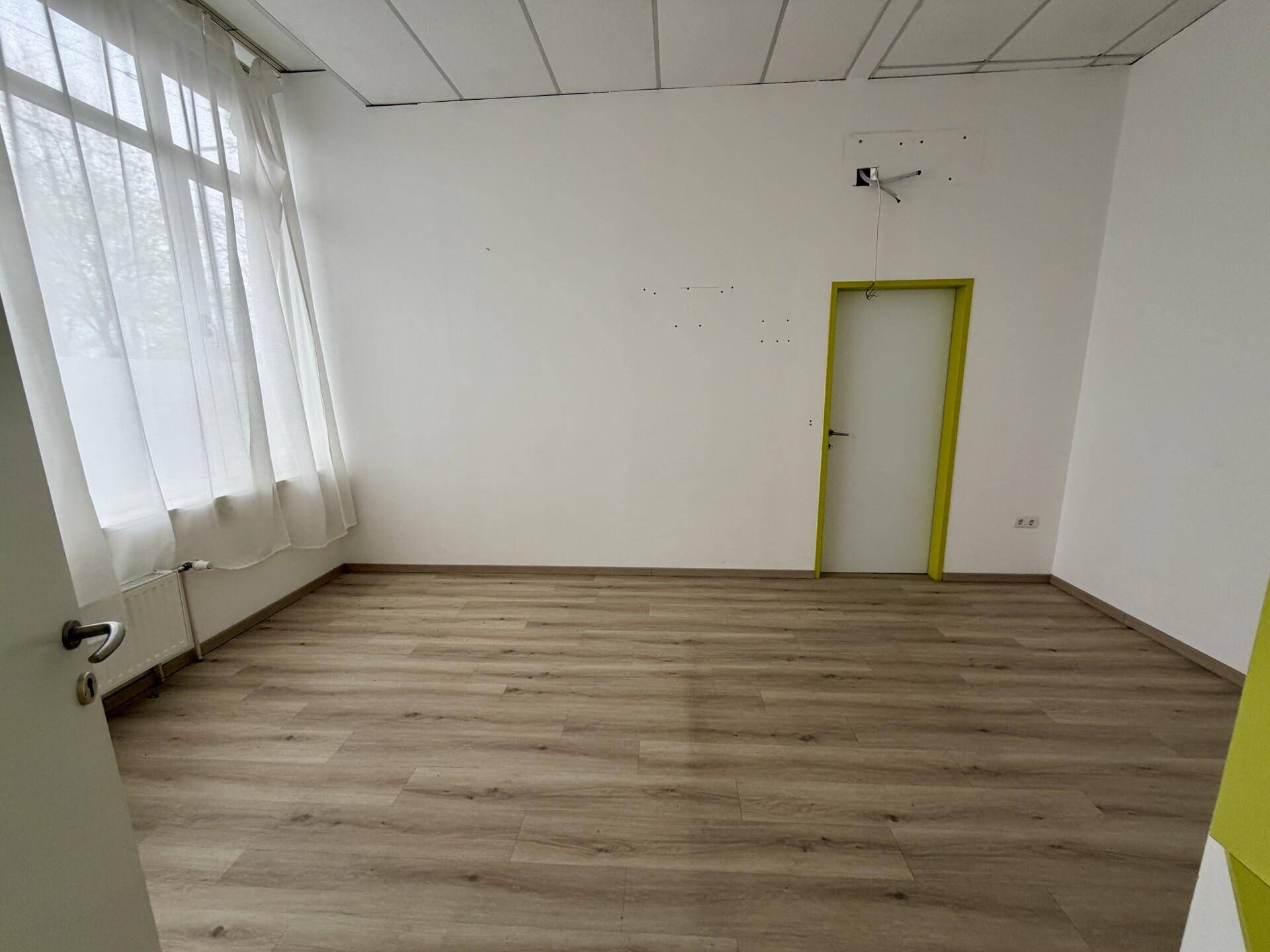Heller Büroraum mit Laminatboden, weißen Wänden und einem Fenster mit Vorhängen.