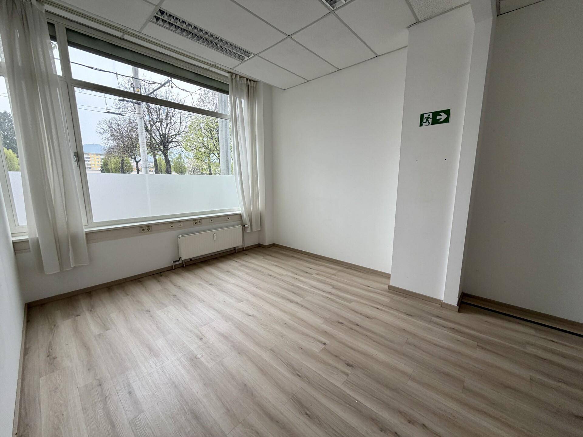 Heller Büroraum mit Laminatboden, großen Fenstern und Vorhängen, die eine angenehme Arbeitsatmosphäre schaffen.