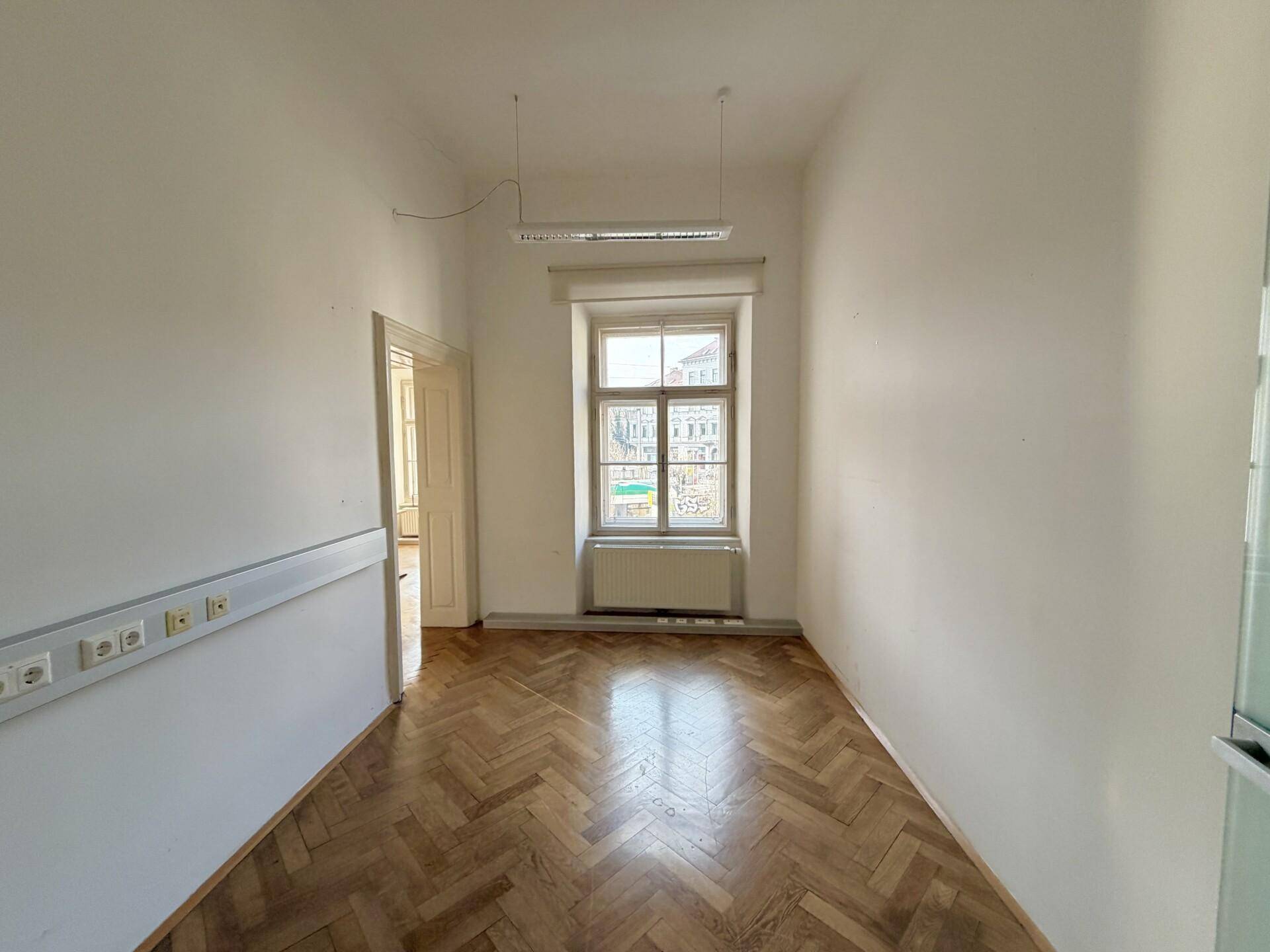 Leeres Büro mit Fischgrätparkett, weißen Wänden und einem großen Fenster, das viel Licht spendet.