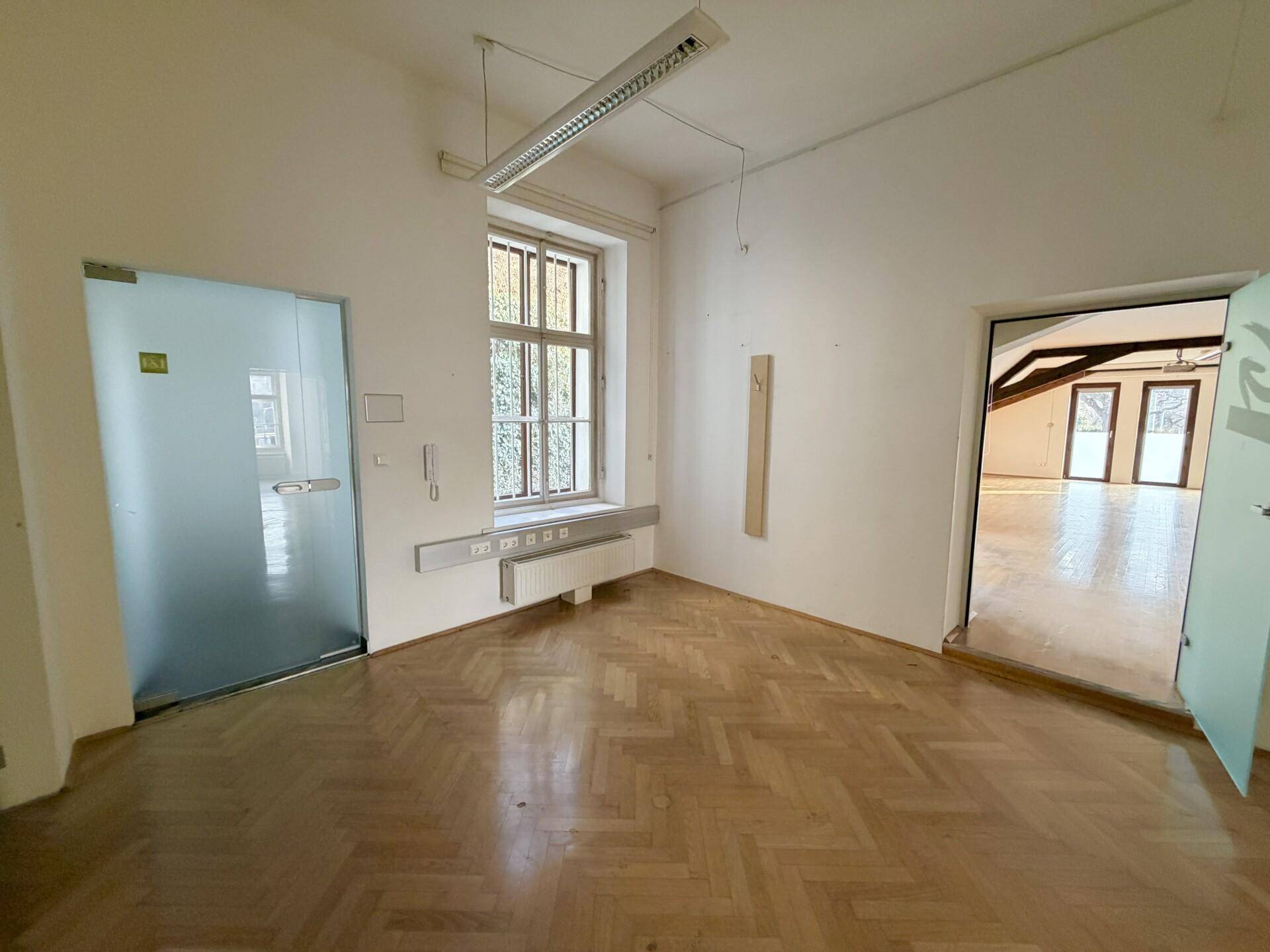 Helles Büro mit Fischgrätparkett, einer modernen Glastür und Blick in einen weiteren Raum.