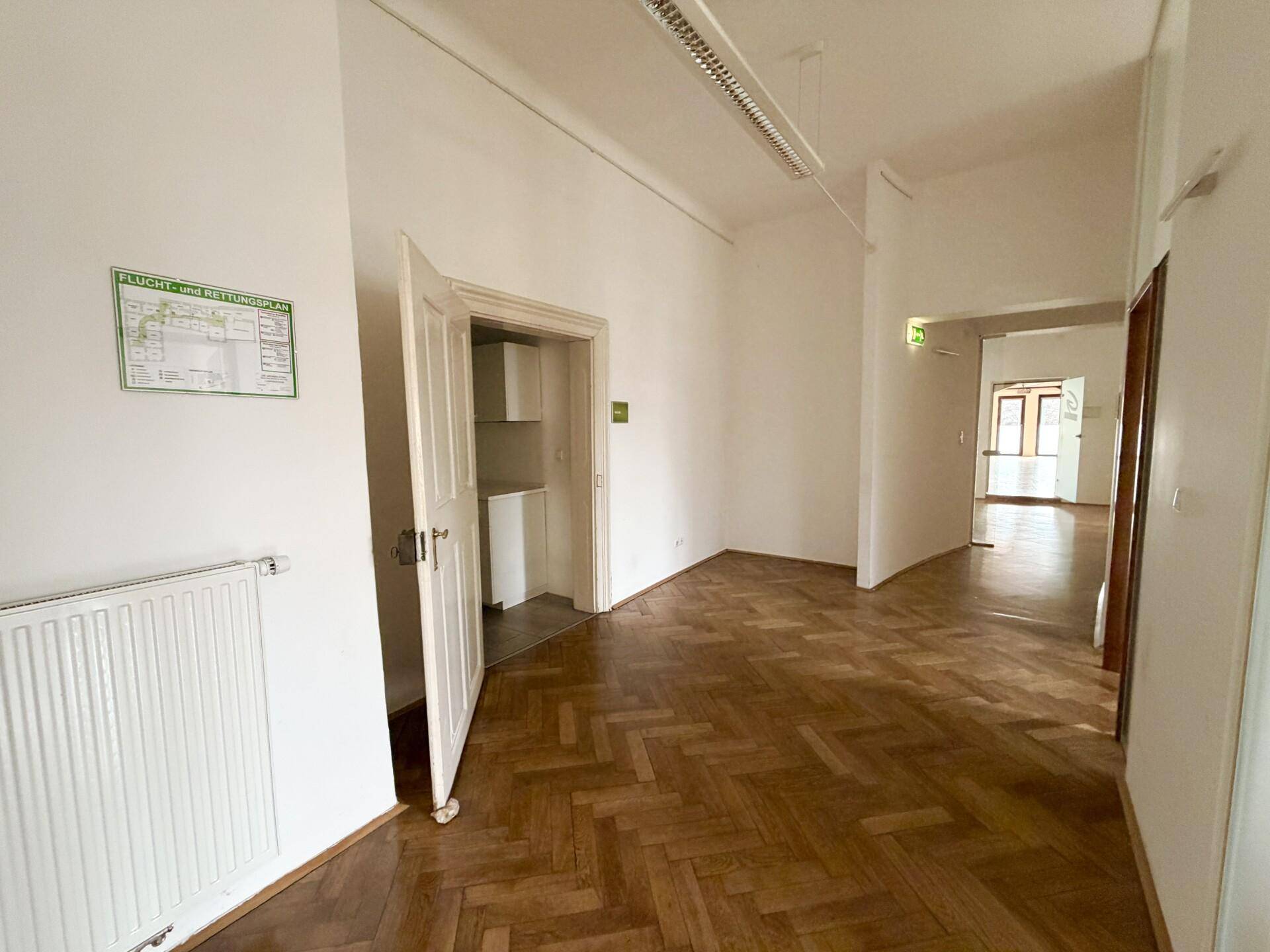 Heller Flur mit Fischgrätparkett, weißen Wänden und einem offenen Durchgang zu weiteren Büroräumen.