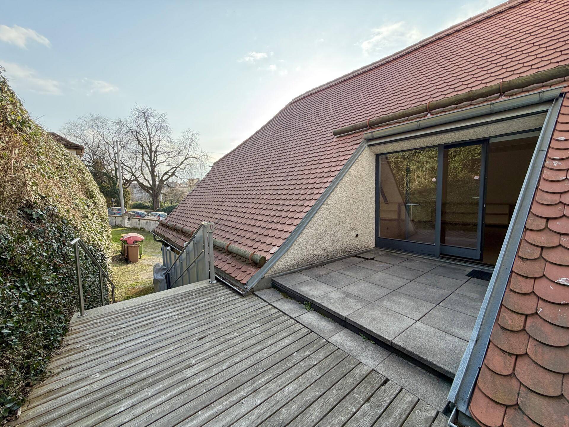 Große Dachterrasse mit Holz- und Fliesenbelag, umgeben von grünen Hecken und Blick auf den Himmel.
