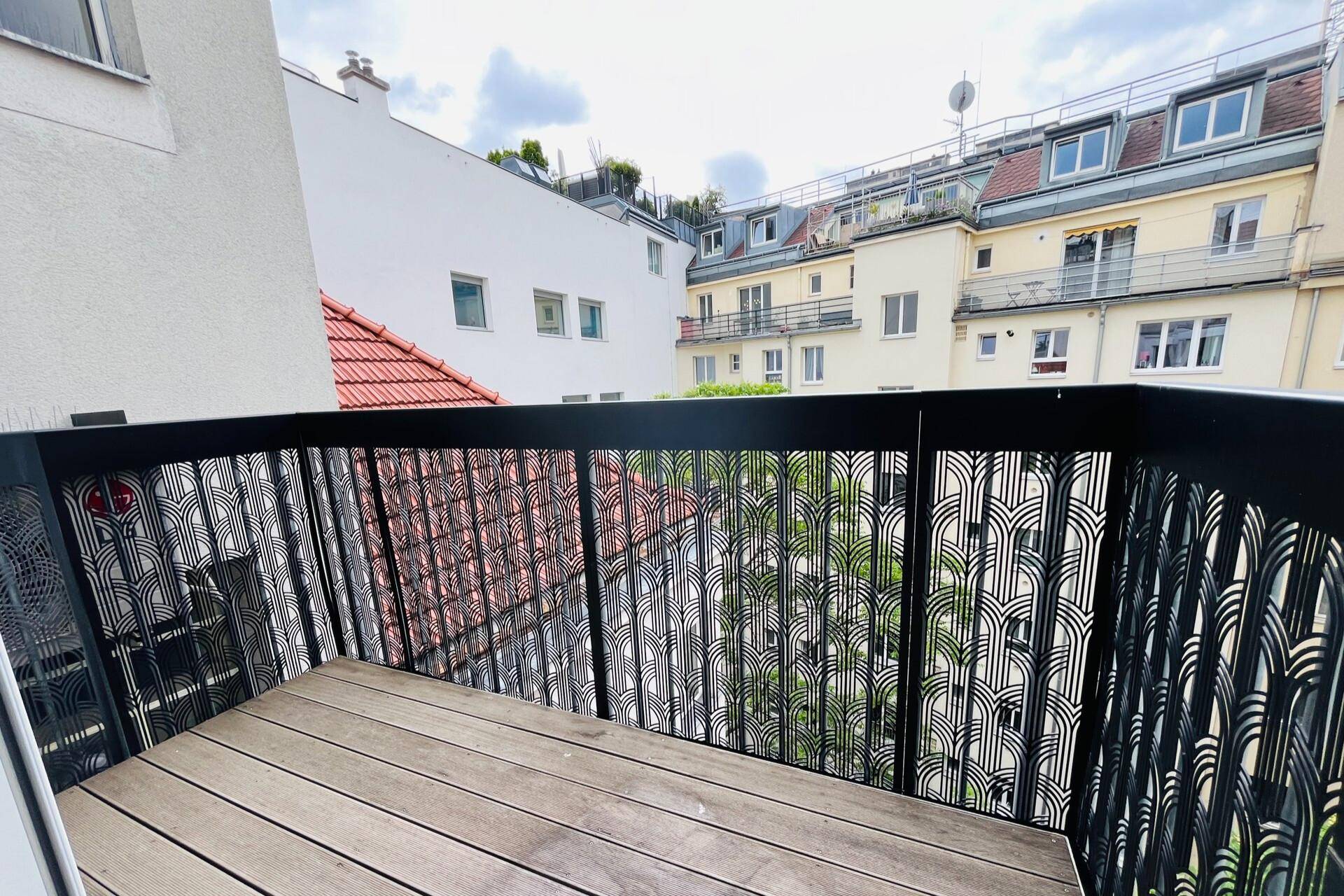 Balkon mit dekorativem schwarzen Geländer und Holzboden, umgeben von städtischen Gebäuden.