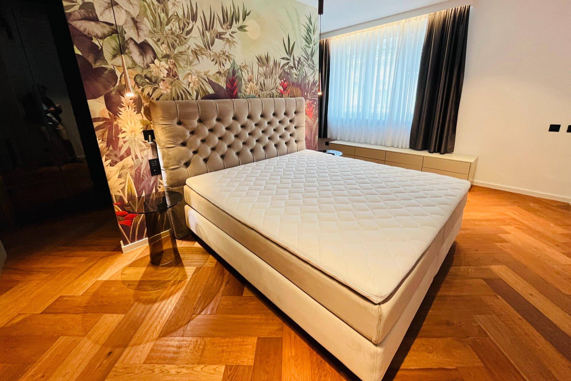 Helles Schlafzimmer mit großem Bett, tropischer Tapete und Holzboden, beleuchtet durch ein Fenster.