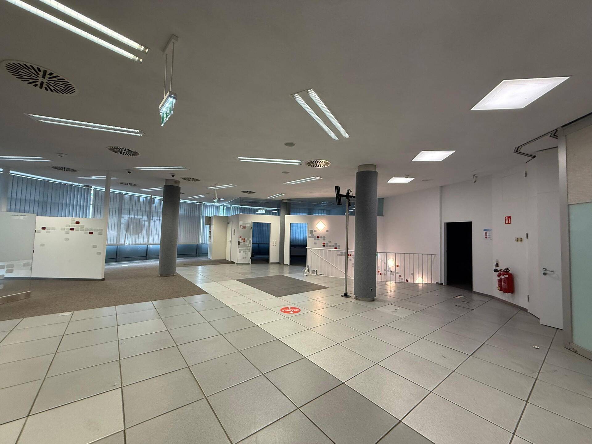 Weitläufiger Büroraum mit verschiedenen Bodenbelägen, Säulen und einem Treppenabgang im Hintergrund.