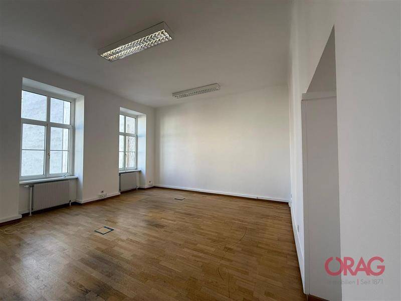 Leerer Büroraum mit hellen Wänden, zwei großen Fenstern und Holzboden.