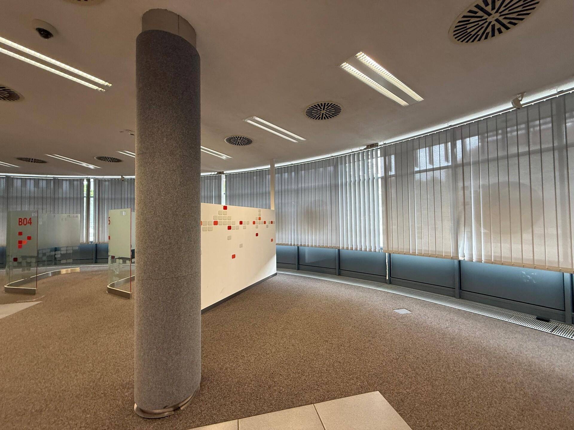 Detailansicht eines Bürobereichs mit Teppichboden, einer Säule und vertikalen Jalousien an den Fenstern.