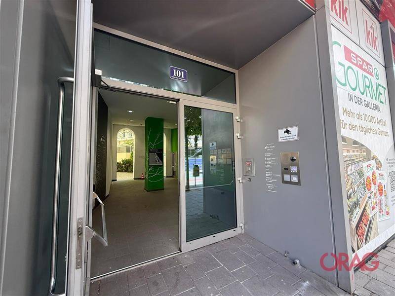 Eingangsbereich eines Gebäudes mit Glastür, Gegensprechanlage und angrenzendem Supermarkt.