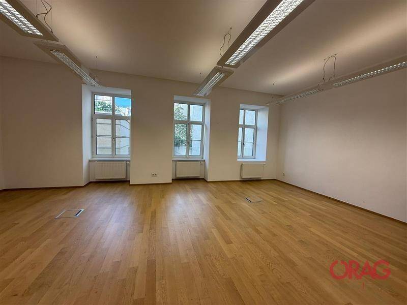Großer, heller Büroraum mit drei Fenstern, Holzfußboden und moderner Deckenbeleuchtung, sehr geräumig.