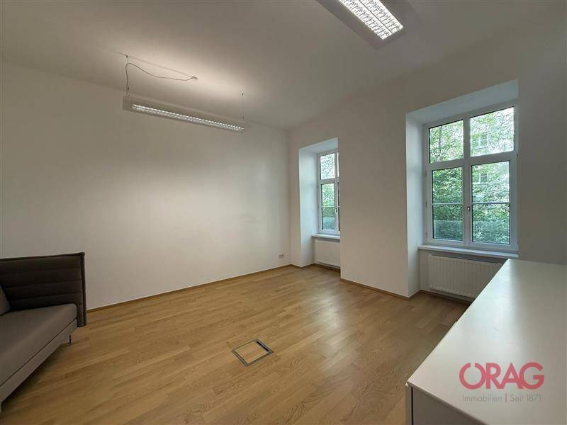 Heller Büroraum mit Holzfußboden, zwei Fenstern und einer kleinen Sitzgelegenheit an der Wand.