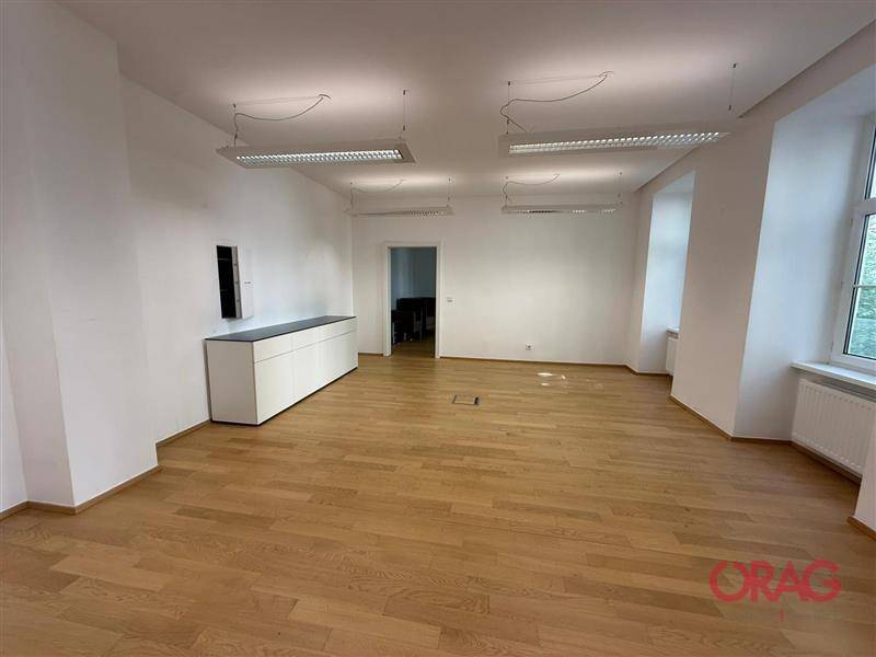 Geräumiger Büroraum mit Holzfußboden, weißen Wänden und Blick auf einen weiteren Raum durch eine Tür.