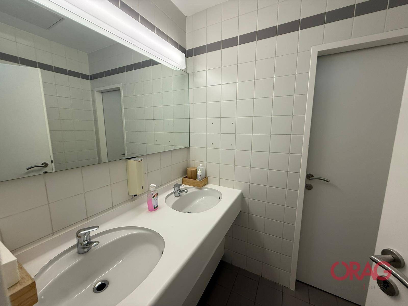 Helles Badezimmer mit zwei Waschbecken, einem großen Spiegel und weißen Fliesen mit grauen Akzentstreifen.
