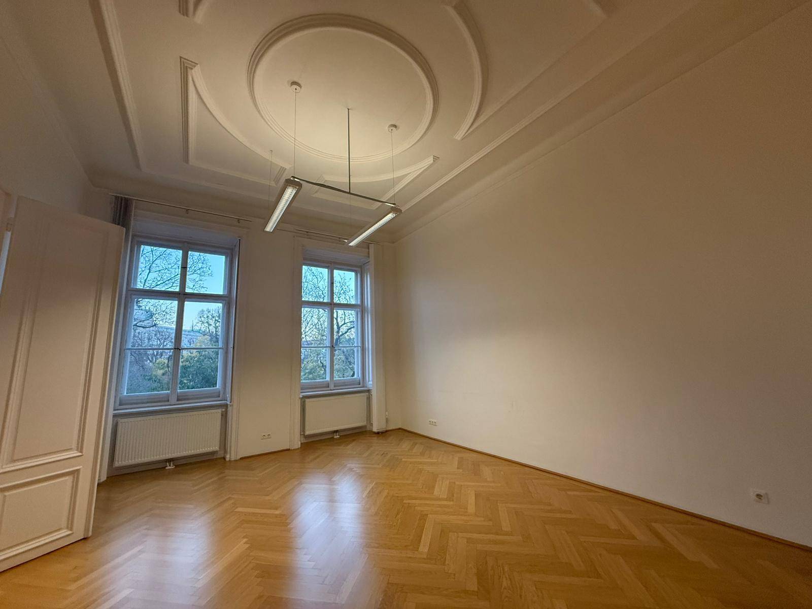 Großer, heller Raum mit Fischgrätparkett, hohen Decken und zwei Fenstern mit Blick nach draußen.