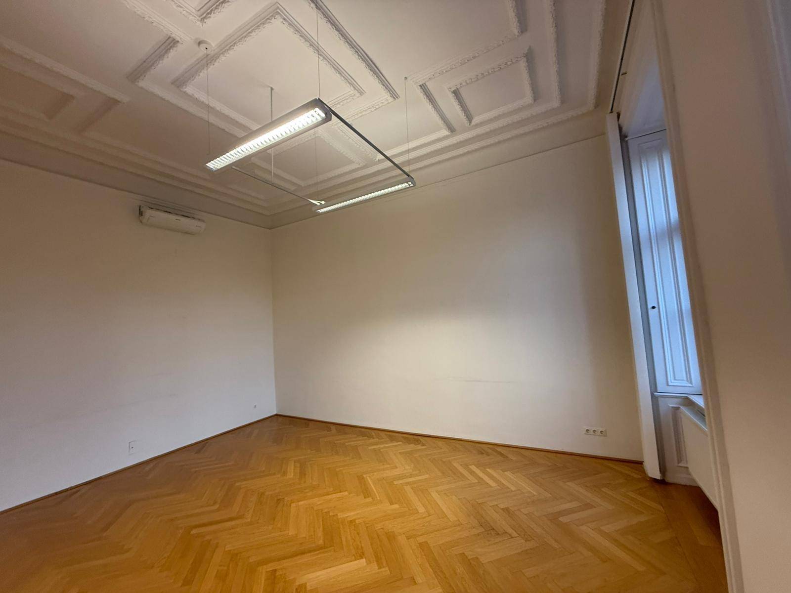 Heller Raum mit Fischgrätparkett, hohen Decken und einem Fenster, das Tageslicht spendet.