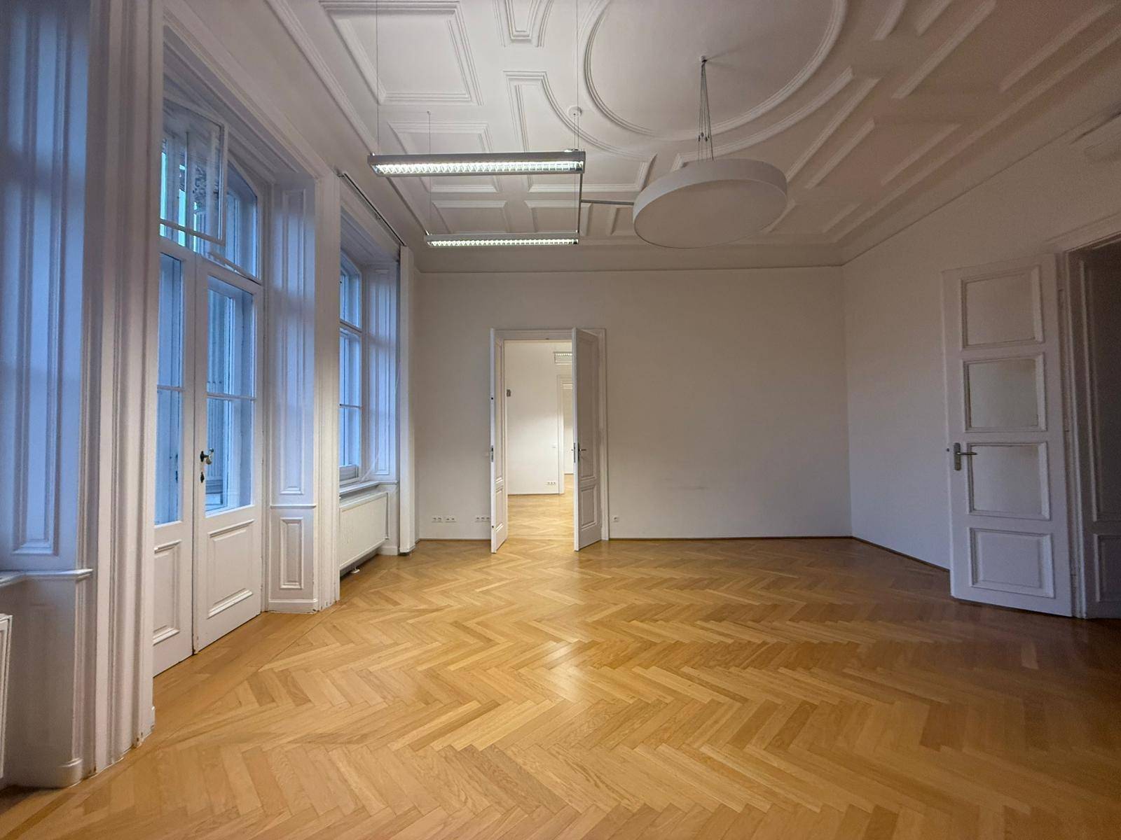 Geräumiger Wohnraum mit Fischgrätparkett, hohen Decken und großen Fenstern, die viel Tageslicht hereinlassen.