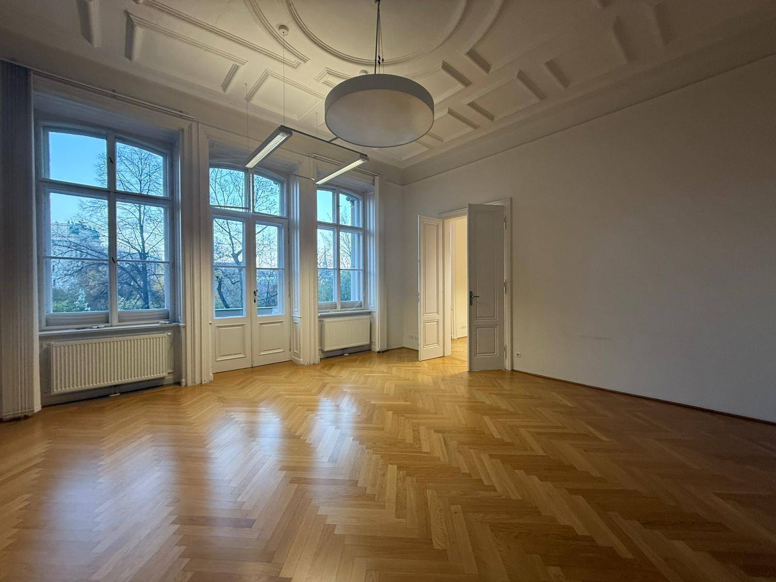 Heller Wohnraum mit klassischem Fischgrätparkett, hohen Decken und Blick ins Grüne durch die Fenster.