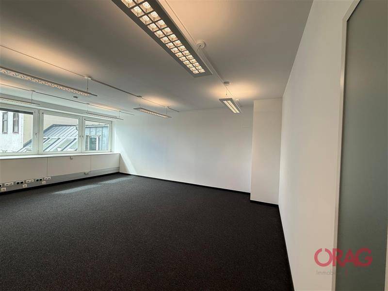 Helles Büro mit Teppichboden, weißen Wänden und großen Fenstern, die viel Tageslicht hereinlassen.