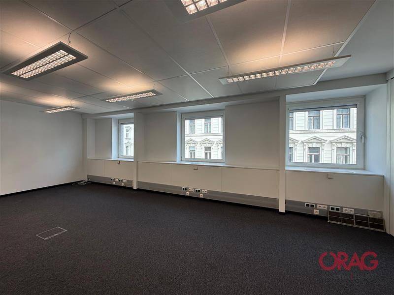 Geräumiges Büro mit Teppichboden, weißen Wänden und mehreren Fenstern mit Blick auf die Stadt.