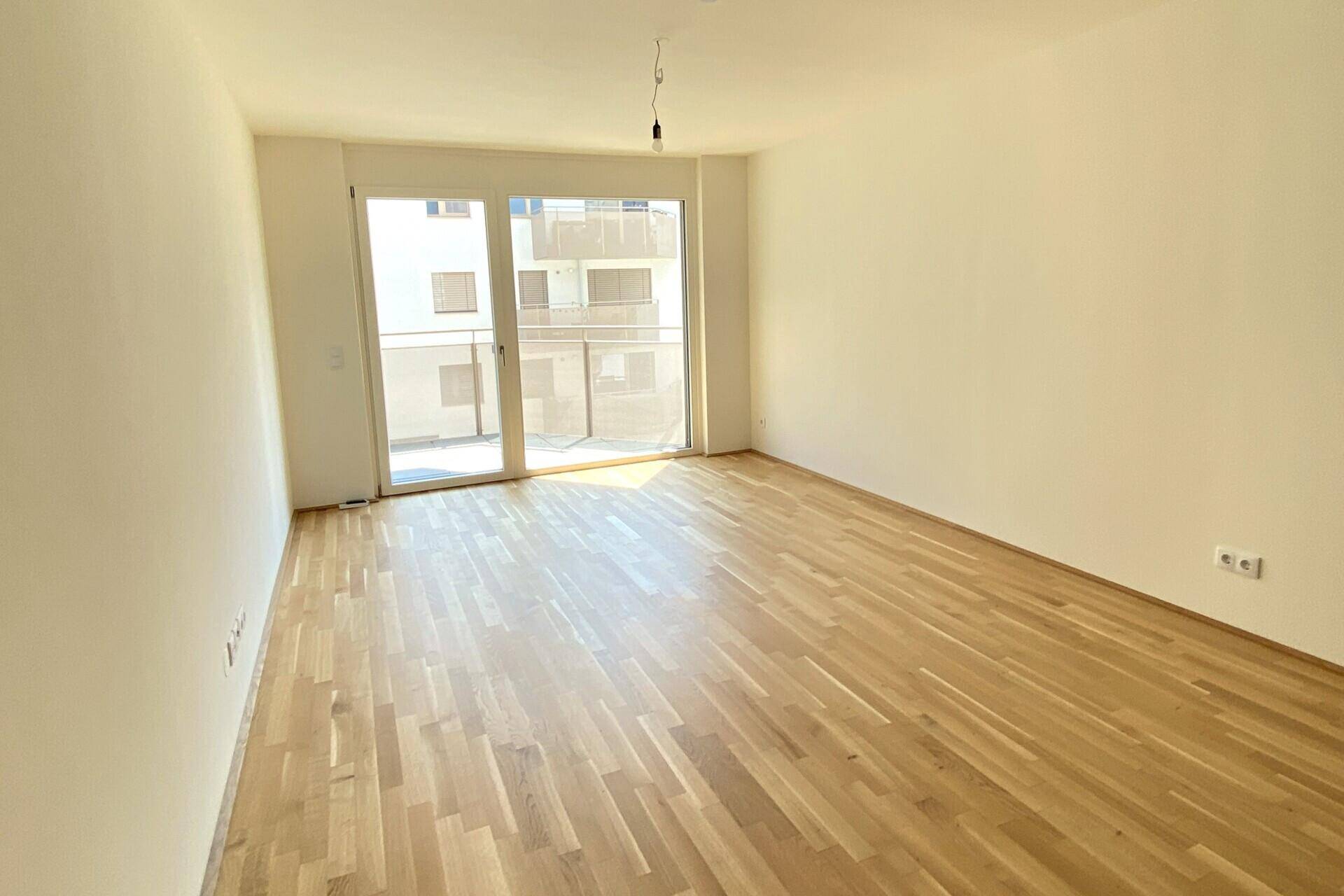 Helles, leeres Wohnzimmer mit Holzboden und Zugang zu einem Balkon.