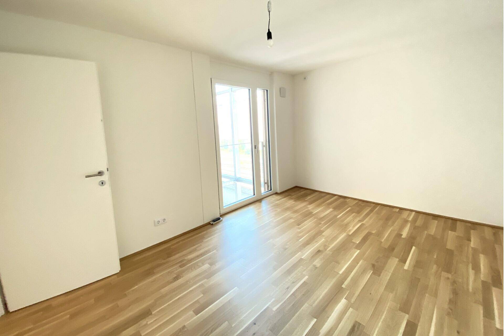 Helles, leeres Schlafzimmer mit Holzboden und Zugang zu einem Balkon.