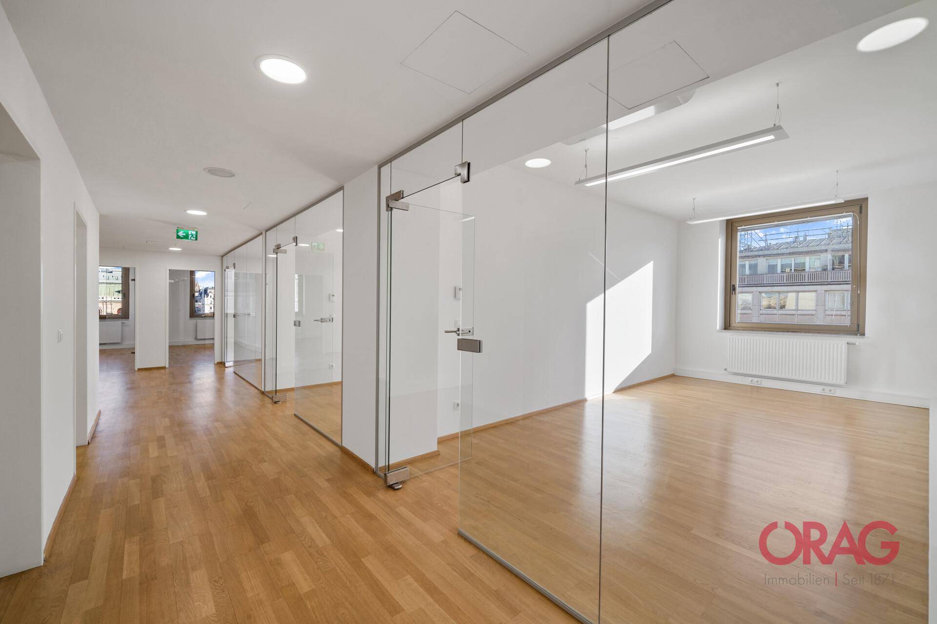 Heller Büroflur mit Glaswänden, die separate Büros abgrenzen, und durchgehendem Holzboden.